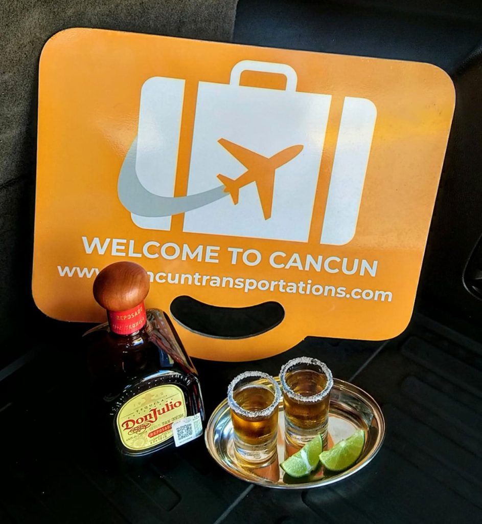 Two served shots of Don Julio tequila in shutter glasses boarded with salt and accompanied by lemons placed over a silver plate in front of Cancun Airport Transportation sign