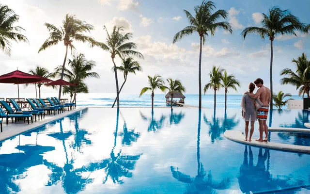 A couple watching the sunrise by the pool of a Playa del Carmen beachfront resort. The image also contains palm trees, a pool, beach chairs, umbrellas a blue sky and a few clouds.