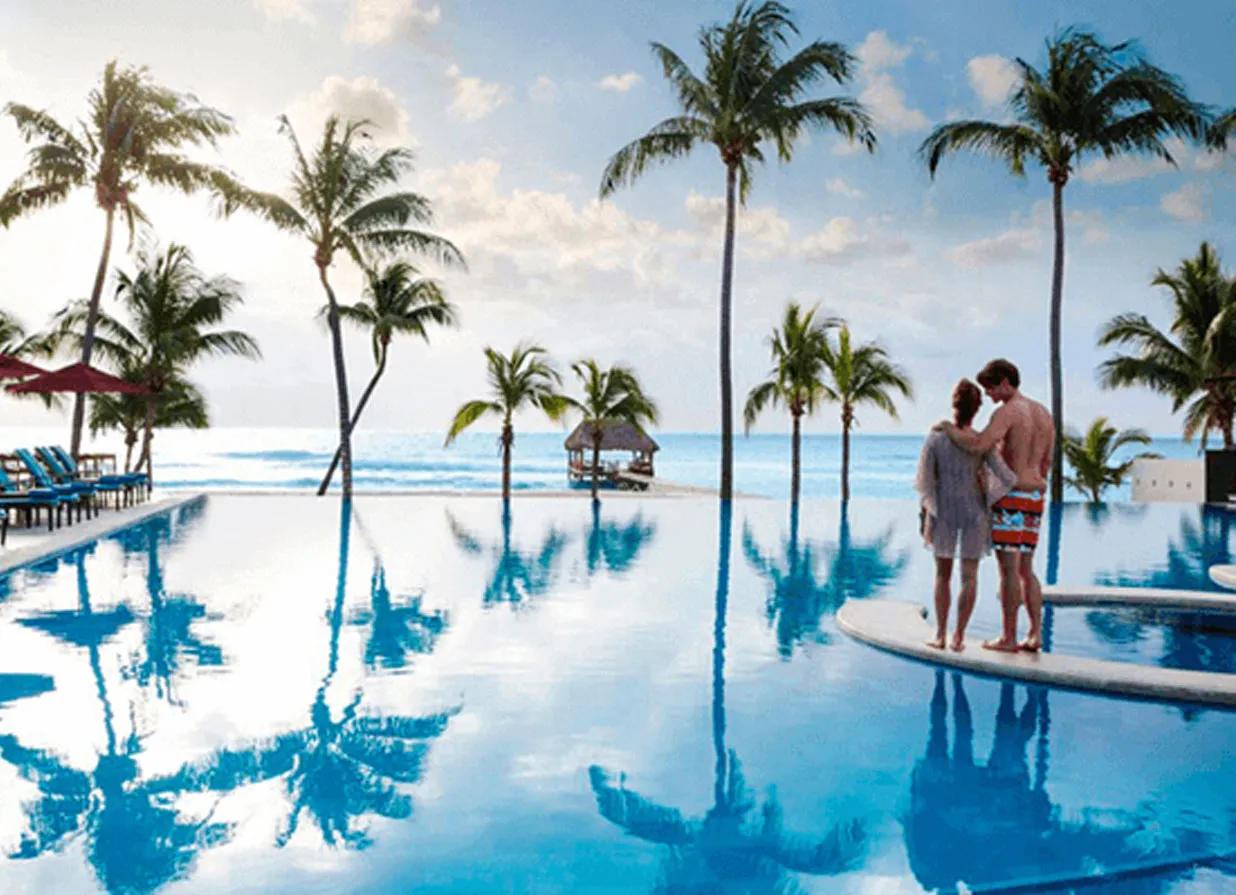 A couple watching the sunrise by the pool of a Playa del Carmen beachfront resort. The image also contains palm trees, a pool, beach chairs, umbrellas a blue sky and a few clouds.